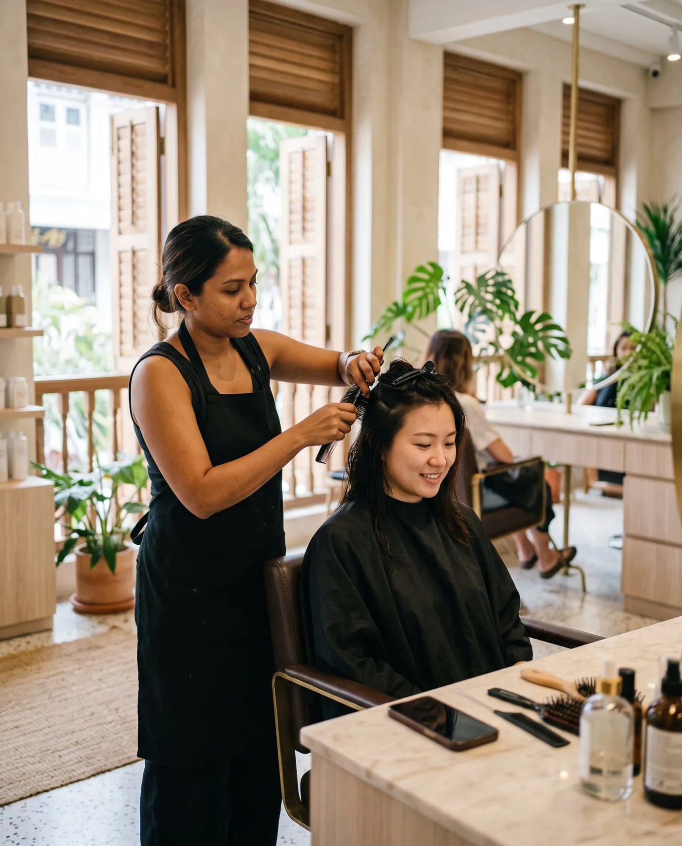 A hairstylist sectioning a client's hair in a bright Singapore shophouse salon, her phone sitting untouched on the counter beside her.